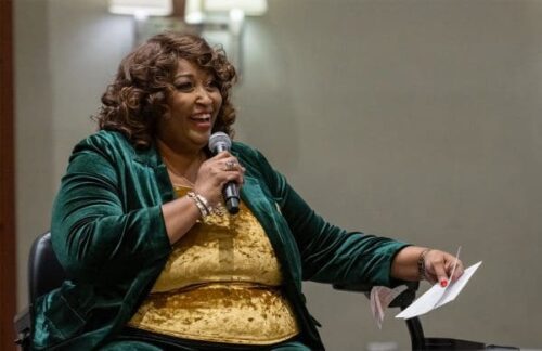 Janine Lee seated in a chair, holding a microphone while presenting at a conference.